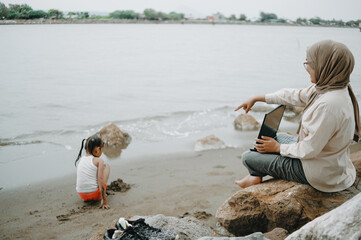 Muslim mother working on laptop on the beach with children playing near the shore
