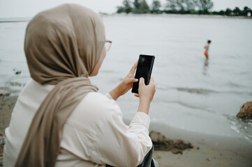 Portrait of a beautiful Muslim woman wearing a hijab using a smartphone on the beach. Technology and communication concept.