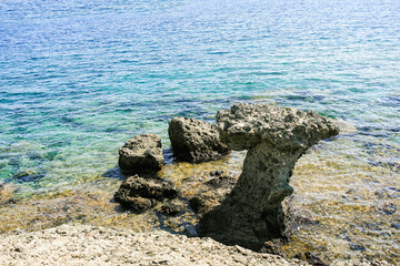 Unique rock formation rises above the clear blue water, surrounded by smaller stones, creating a serene coastal scene that captures the beauty of nature and geological wonders