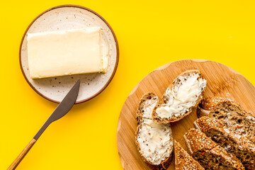 Wholegrain bread on cutting board with butter on plate, slice of bread with butter on yellow background top view