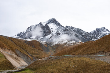 Majestic Himalayan peaks dusted with snow rise above rugged brown hills. This dramatic, overcast...