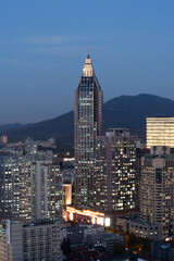 Naklejka premium A striking, uniquely designed skyscraper illuminates the city skyline at blue hour. This high-angle view captures vibrant urban lights set against the dark silhouette of a mountain.