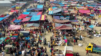 Bogura, Bangladesh - 17 August 2025: Aerial view of a bustling market scene filled with colorful stalls and crowds of people moving along the sandy ground.