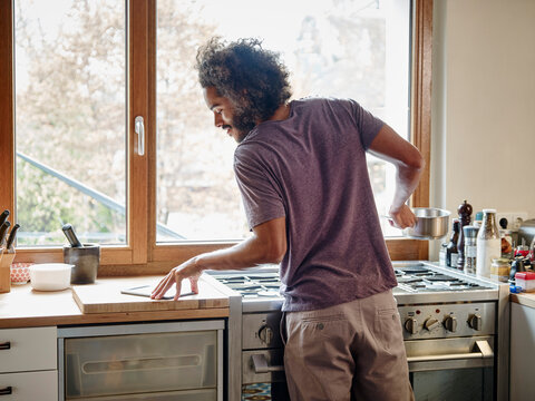Young man standing at stove in kitchen checking digital tablet