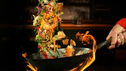 Freeze Motion of Wok Pan with Flying Ingredients in the Air and Fire Flames. Kitchen Interior on Background.