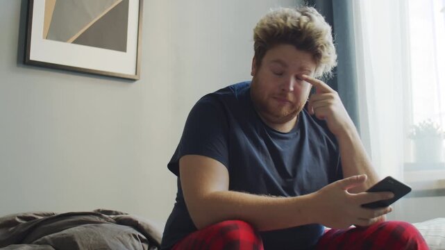 Horizontal low angle view shot footage of young Caucasian man sitting on bed in morning rubbing eyes and watching something on smartphone