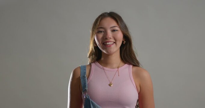 Studio video clip of a young adult female making different expressions, both positive and negative, towards the camera. She has long brunette hair and is wearing denim dungarees with a pale pink top. 