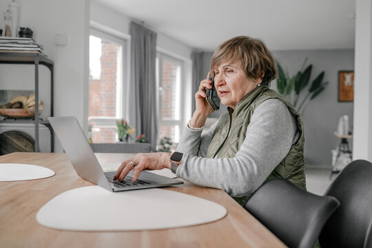 Elderly woman using laptop and talking on smart phone at home
