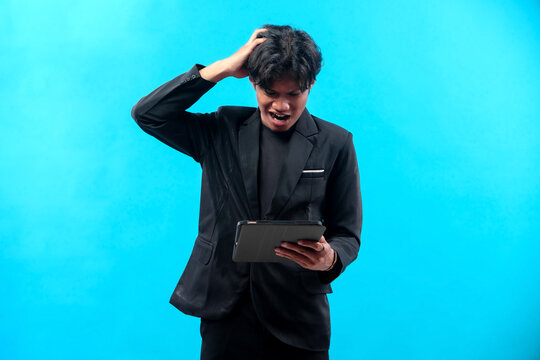 A young man in a black suit scratching his head while looking at a tablet with a frustrated expression, symbolizing stress, confusion, business problems, and digital challenges.