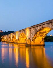 Illuminated stone bridge spanning a calm river at twilight, reflecting lights in the water