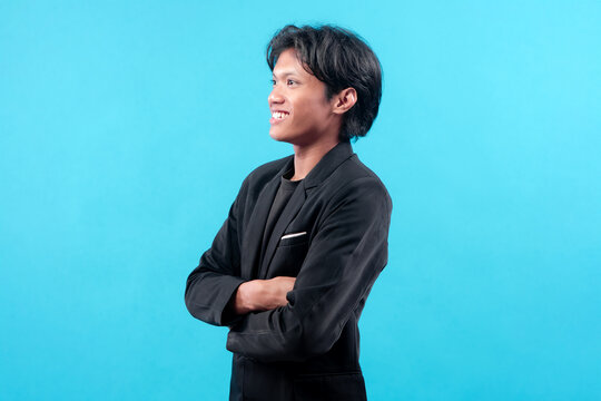 A young man in a black suit standing in side profile with folded arms and a cheerful smile, representing confidence, optimism, and modern professional style on blue background.