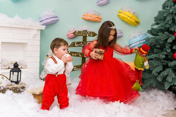 A little girl and a boy in red clothes have fun in the artificial snow by the fireplace in the studio. The girl is holding a box with gifts. Little kids in red christmas costume having fun beside a