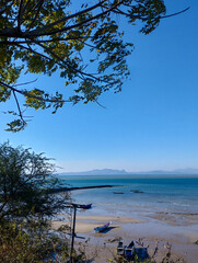 Scenic coastal view with small fishing boats on a sandy shore, calm turquoise sea, and distant mountains under a clear blue sky