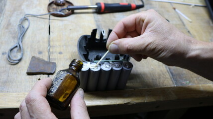 Processing contacts on old batteries from a construction tool during device resuscitation, close-up of male hands applying solution from a brown glass bottle to battery contacts on a work table