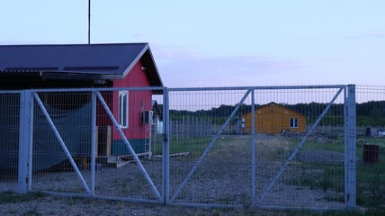 Red farmhouse behind white metal gates and orange building in the distance, farm provincial landscape with residential and outbuildings, no people, farmer's housing in the countryside