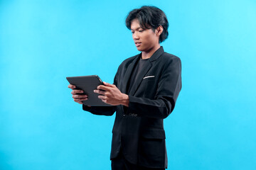 A focused young Asian man in a formal black suit is holding a digital tablet, appearing concentrated and professional while engaging with modern technology against a blue background.