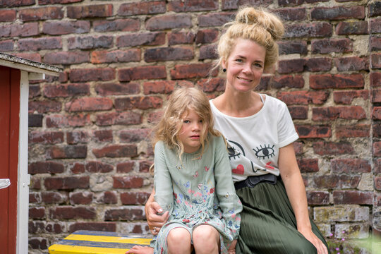 Smiling woman with daughter with blond hair sitting against brick wall at back yard