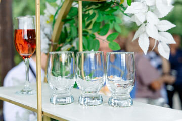 Four empty glasses, one of which is filled with a red drink, are neatly arranged on the table. The background is decorated with greenery and white flowers, creating a festive atmosphere.
