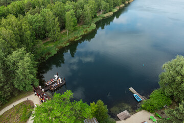 Aerial view of a wedding celebration by the lake, guests gathered on a wooden pier and terrace surrounded by lush green forest and a modern glass building in summer time.
