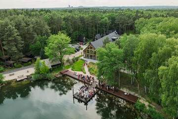 Aerial view of a wedding celebration by the lake, guests gathered on a wooden pier and terrace surrounded by lush green forest and a modern glass building in summer time.