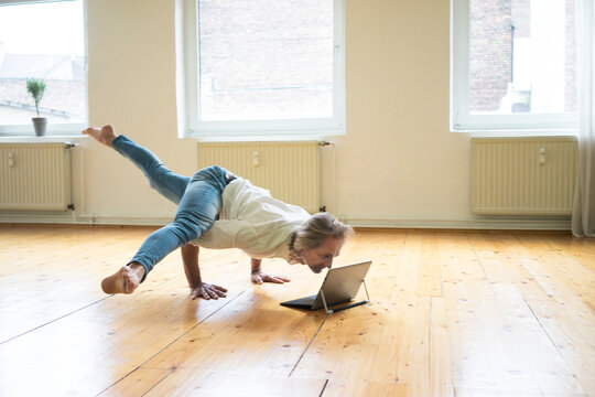 Mature man doing a handstand on floor in empty room looking at tablet