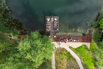 Aerial view of a wedding celebration by the lake, guests gathered on a wooden pier and terrace surrounded by lush green forest and a modern glass building in summer time.
