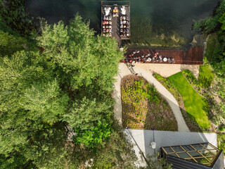 Aerial view of a wedding celebration by the lake, guests gathered on a wooden pier and terrace surrounded by lush green forest and a modern glass building in summer time.