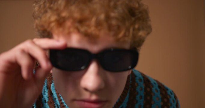  Video clip of a young adult male wearing sunglasses in the studio, walking close to the camera to check his appearance. He has curly hair and is wearing casual clothing.