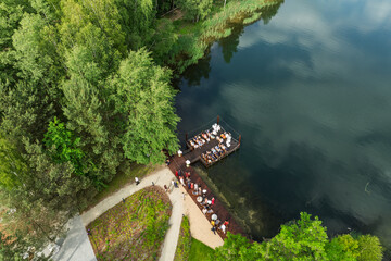 Aerial view of a wedding celebration by the lake, guests gathered on a wooden pier and terrace surrounded by lush green forest and a modern glass building in summer time.