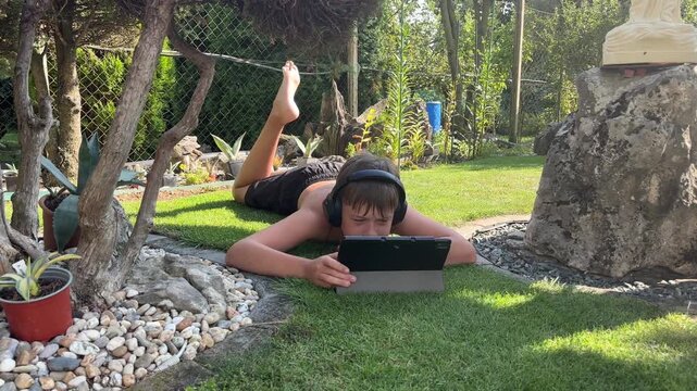 A boy is lying on a green lawn in a backyard, using a tablet with headphones on, enjoying a relaxing summer day in a comfortable home setting, of home entertainment, children, summer activities - Powered by Adobe