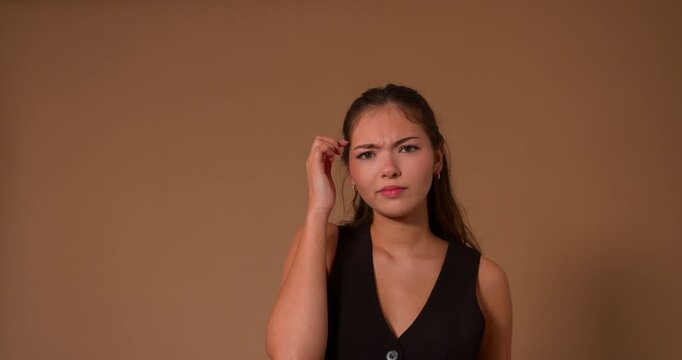 Studio video clip of a young adult female making puzzled expressions and shrugging her shoulders towards the camera. She has long brunette hair and is wearing smart casual clothing.