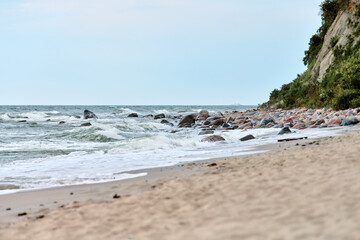 Dynamic waves crash against rocky shoreline under soft daylight. Sandy beach foreground contrasts with textured cliffside. Natural baltic beauty in serene coastal landscape