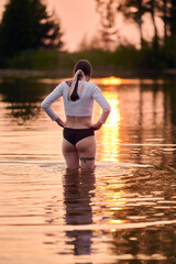 Fototapeta premium Young woman with brown hair stands waist-deep in water, admiring sunset. Warm orange hues create serene atmosphere, reflecting on calm surface. Trees silhouette in background