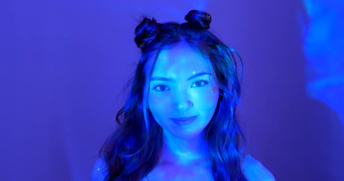  Video clip of a young adult female smiling in the studio. She has long brunette hair and she is illuminated by blue neon lights.