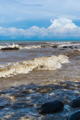 Beautiful black-brown waves crashing on the rocks at Celong Beach, Batang, Indonesia, under a stunning blue sky with white clouds