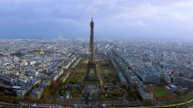4K drone view of Paris with the Eiffel Tower centered over Champ de Mars, flanked by Montparnasse Tower and Les Invalides - dramatic winter cityscape under moody clouds.