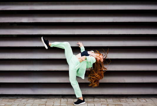 Redhead woman bending backwards with leg up dancing on footpath near wall