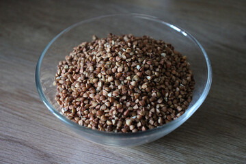 Horizontal photo of a clear glass bowl with dry buckwheat grains on wooden surface, natural organic product used for healthy meals