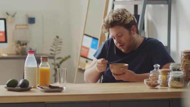 Young Caucasian man spending morning at home eating oats with milk for breakfast and chatting on phone with someone