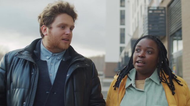 Young Caucasian man and Black woman walking along street on autumn day talking about something