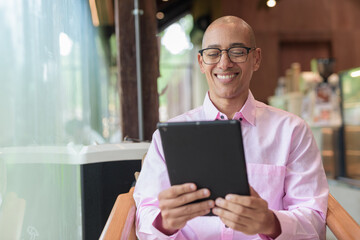 Bald mature Hispanic man wearing pink casual business shirt in coffee shop using tablet computer