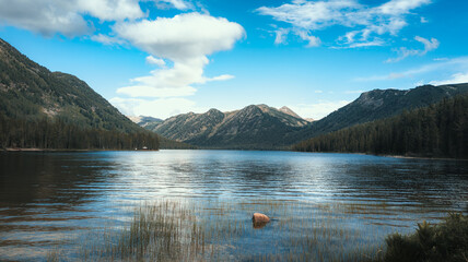 A serene mountain lake reflects greenery and clouds, evoking tranquility. Peaceful beauty of unspoiled nature portrayed.