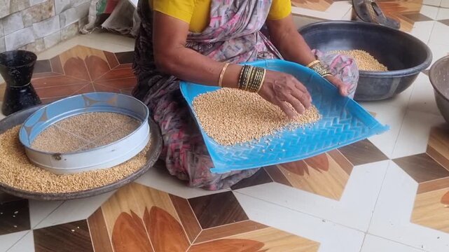 An Indian woman engaged in the traditional agricultural process of winnowing Jowar (sorghum). The woman is using a winnowing basket (likely a muram in India) to separate the lighter chaff.