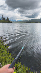 A person with a fishing rod by a calm lake under stormy skies, highlighting the connection between nature and human presence.