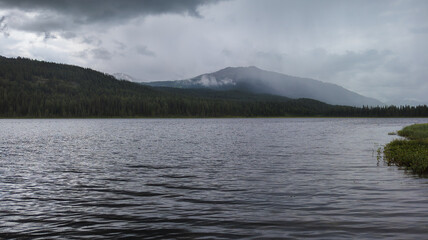 Tranquil image of a calm lake surrounded by trees and mountains with mist. Overcast sky reflecting on the water creates a serene, peaceful atmosphere.