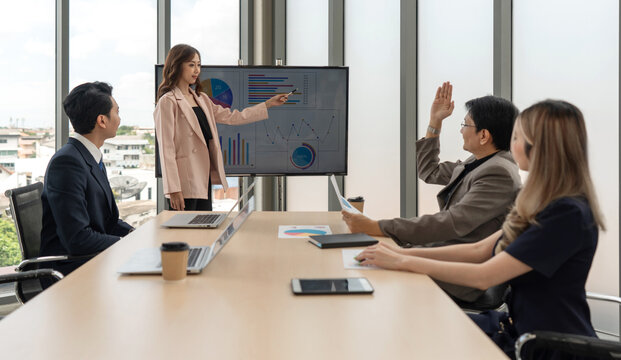 Asian businessman in suit raise his hand to ask a question to presenter standing in front of a large digital monitor. Business executives team meeting in modern office.