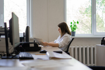 Woman in white shirt sitting at computer with paper by window