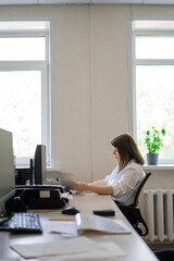 Woman in white shirt sitting at computer with paper by window