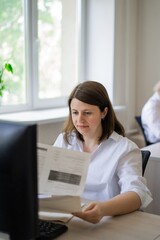 Woman in white shirt sitting at computer with paper by window