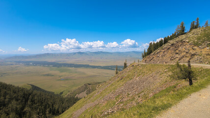 A winding road cuts through mountains, rocks, and pine trees in a vast landscape under a clear blue sky with scattered clouds.
